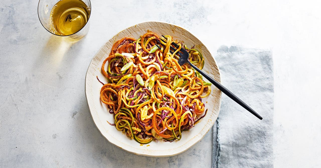 A plate of spiralized vegetables, including carrots and zucchini, garnished with sliced green onions, next to a glass of light-coloured drink.