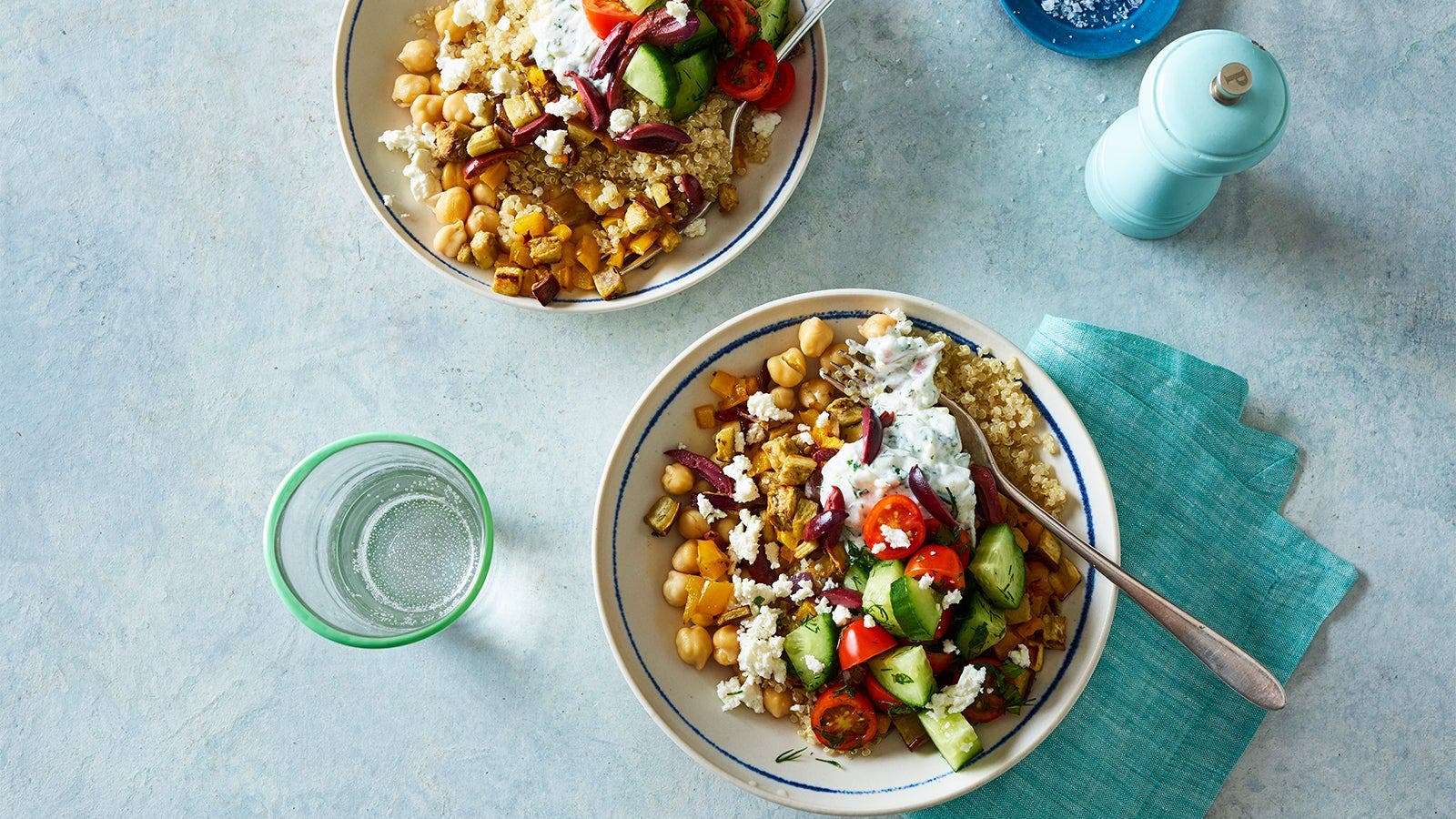 Two bowls of a vibrant quinoa salad topped with chickpeas, feta cheese, chopped vegetables, and tzatziki sauce, set on a light blue surface with a glass of water.