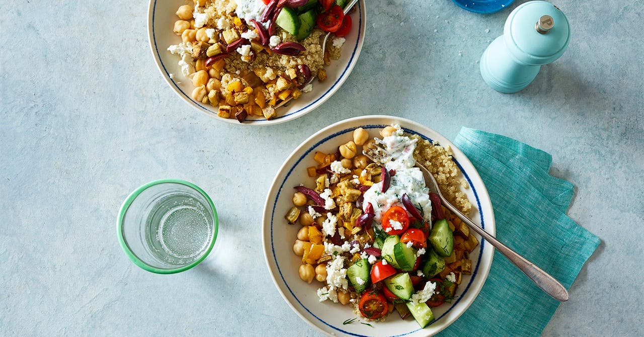 Two bowls of a vibrant quinoa salad topped with chickpeas, feta cheese, chopped vegetables, and tzatziki sauce, set on a light blue surface with a glass of water.