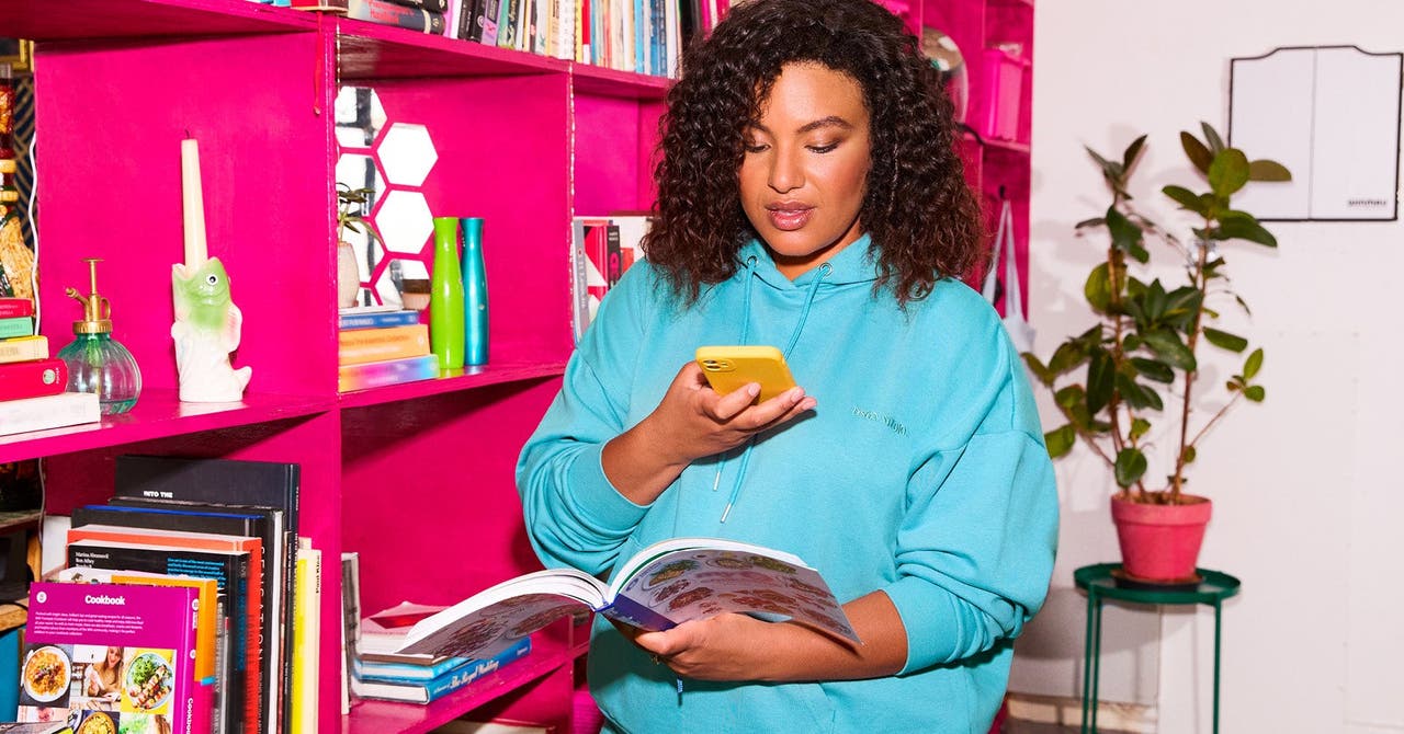 Woman in a turquoise hoodie reading a cookbook while holding a yellow smartphone by a pink bookshelf.