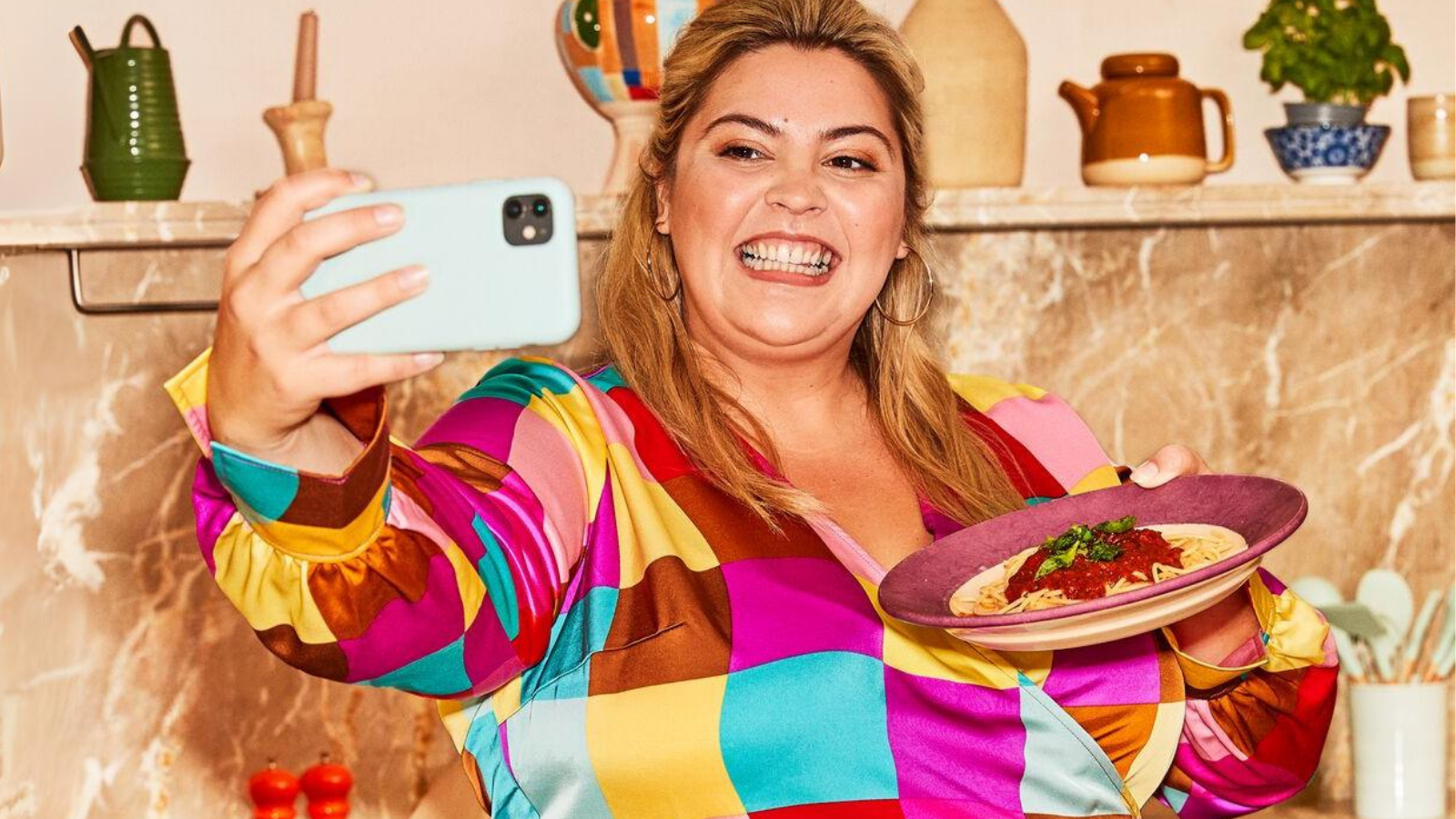 A woman in a colorful shirt smiling and taking a selfie while holding a plate of pasta in a kitchen.