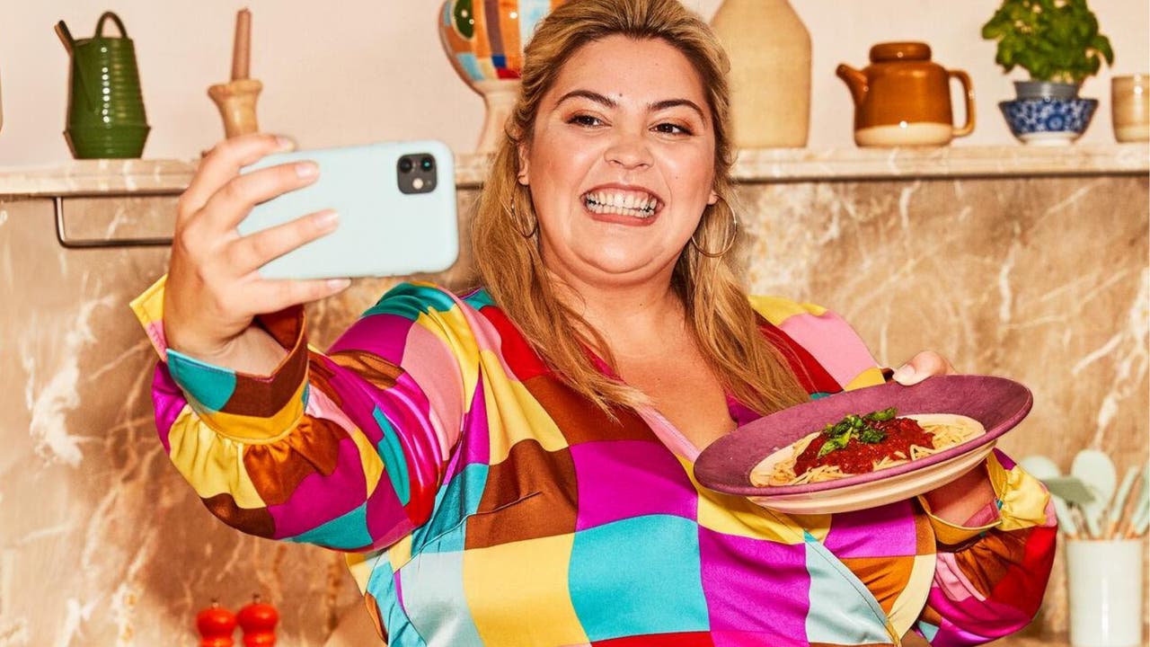 A woman in a colorful shirt smiling and taking a selfie while holding a plate of pasta in a kitchen.