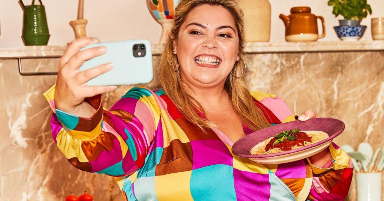 A woman in a colorful shirt smiling and taking a selfie while holding a plate of pasta in a kitchen.