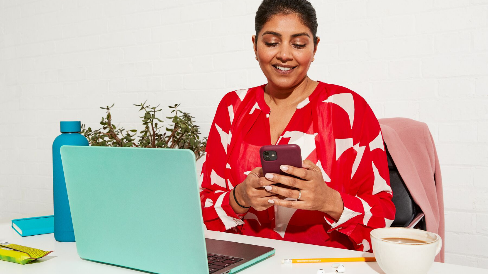 A woman sitting at a desk using a smartphone, with a laptop, coffee, and snacks nearby.