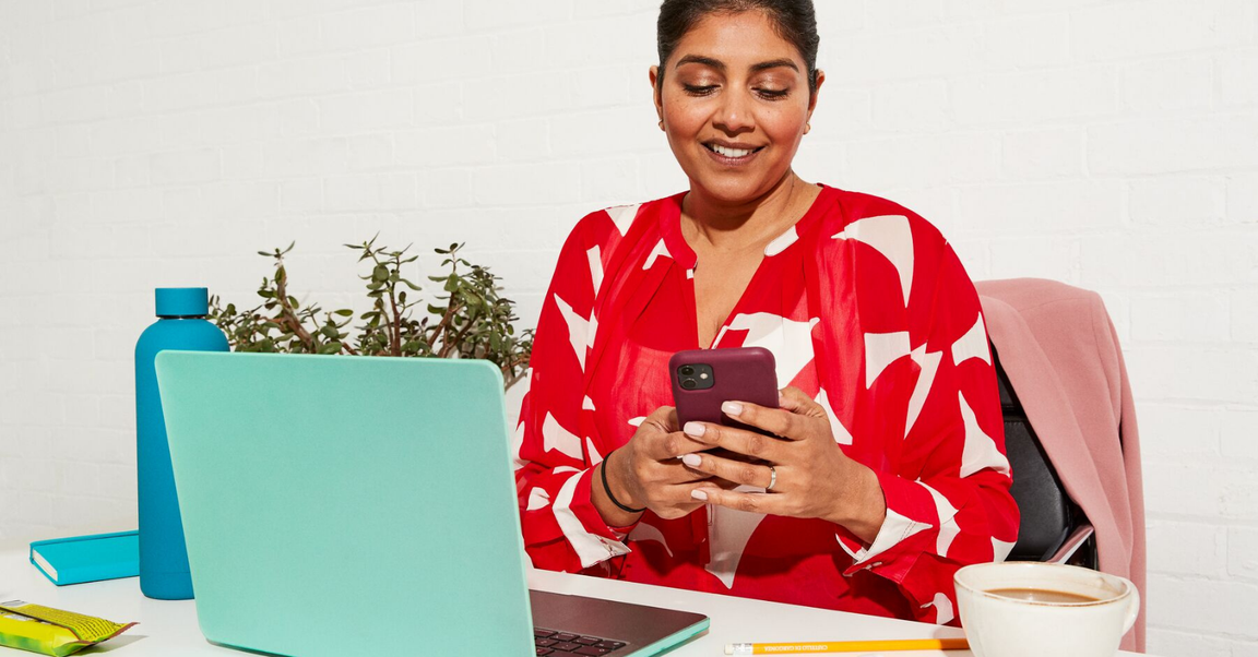 A woman sitting at a desk using a smartphone, with a laptop, coffee, and snacks nearby.