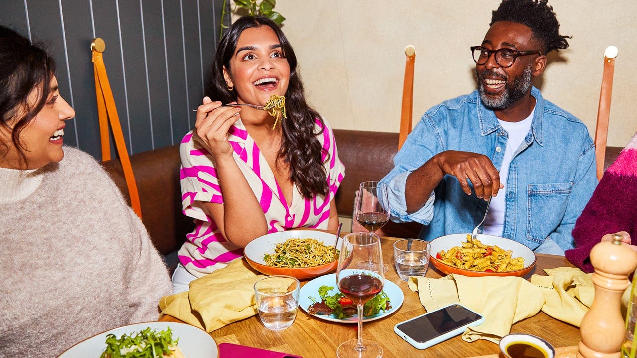 A group of friends enjoying a meal together at a restaurant, with one woman laughing and holding a fork with pasta.