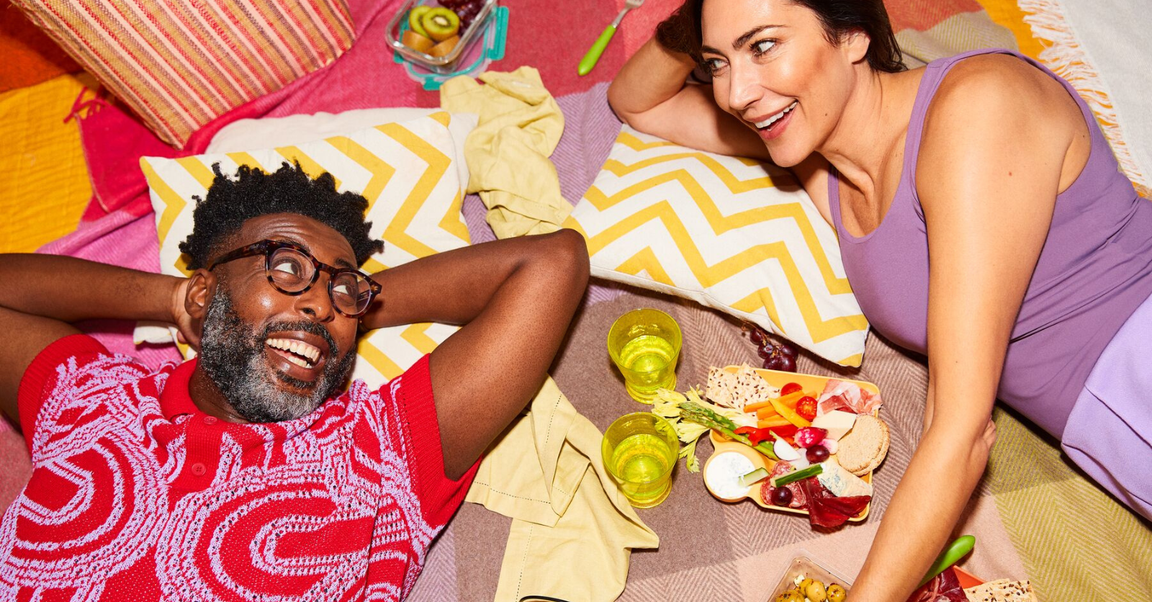 A couple relaxes on a picnic blanket, enjoying snacks and drinks, with the man wearing a red shirt and the woman in a purple top.