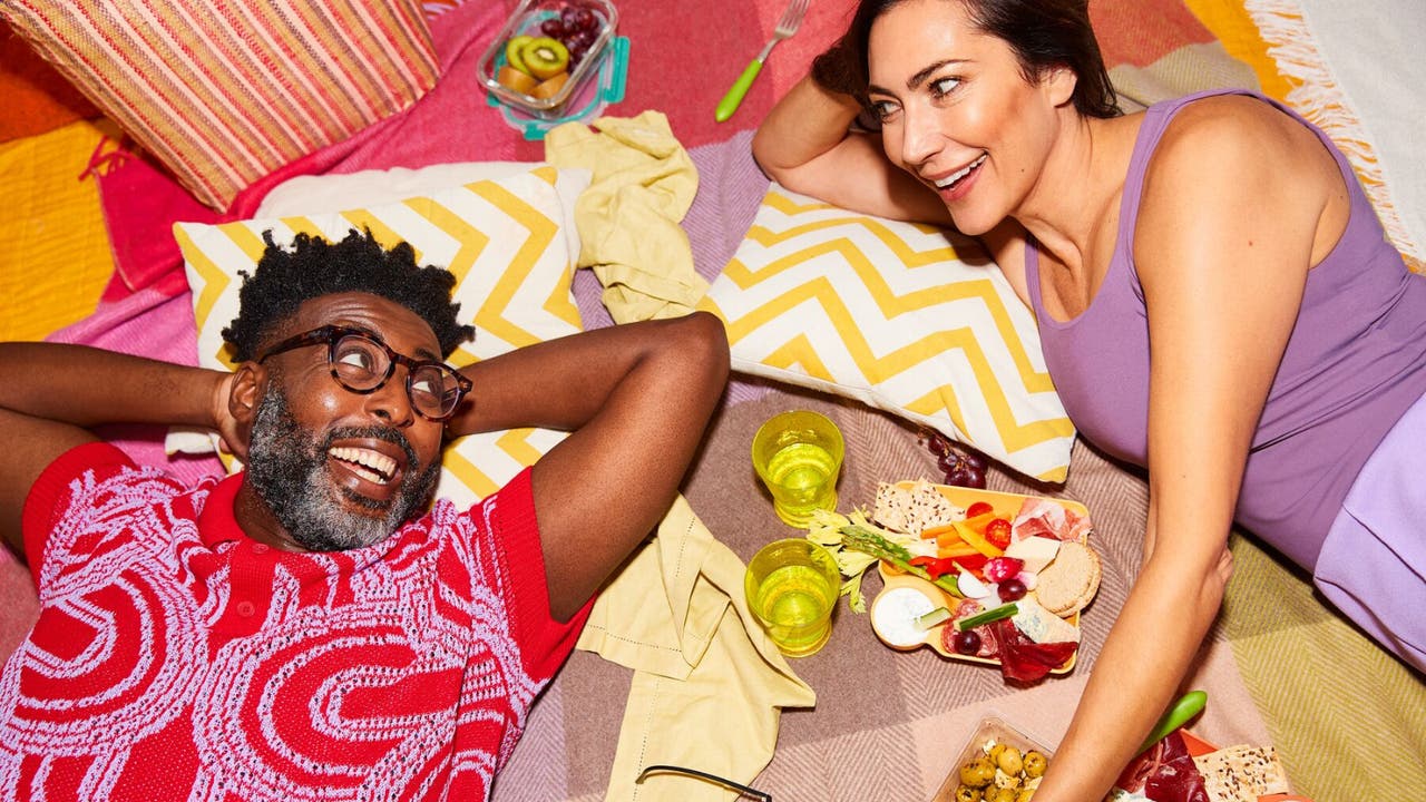 A couple relaxes on a picnic blanket, enjoying snacks and drinks, with the man wearing a red shirt and the woman in a purple top.
