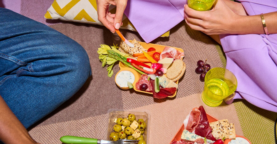 A close-up view of a picnic setting with two people enjoying a colourful platter of snacks, including vegetables, cheese, and cured meats