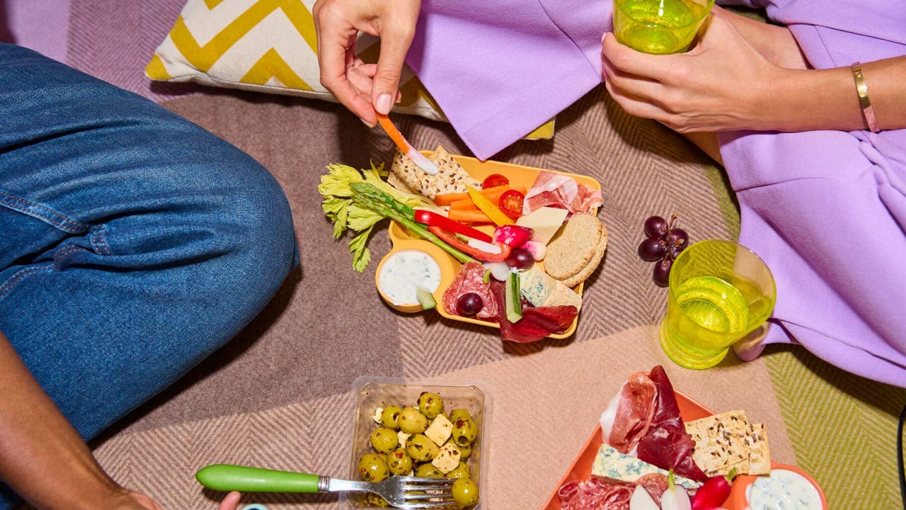 A close-up view of a picnic setting with two people enjoying a colourful platter of snacks, including vegetables, cheese, and cured meats