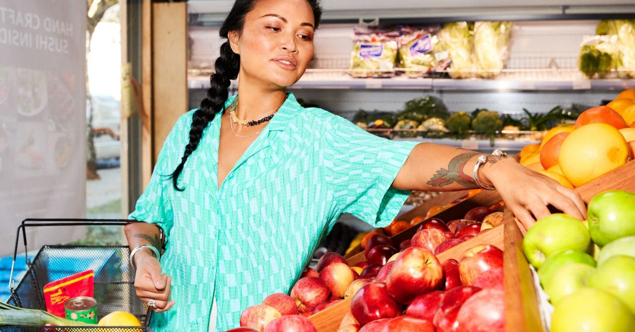 Woman selecting apples in a grocery store, wearing a green patterned shirt.