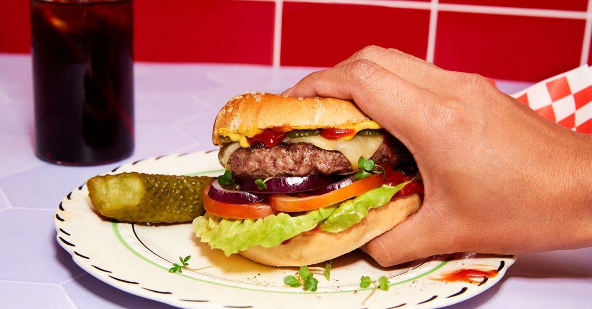 Close-up of a hand holding a burger with lettuce, tomato, cheese, and pickles, with a pickle on the side.