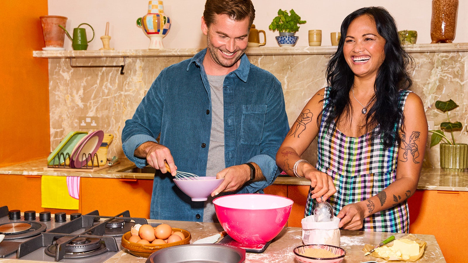 A man and a woman laughing while baking together in a kitchen, surrounded by baking ingredients like eggs and flour.