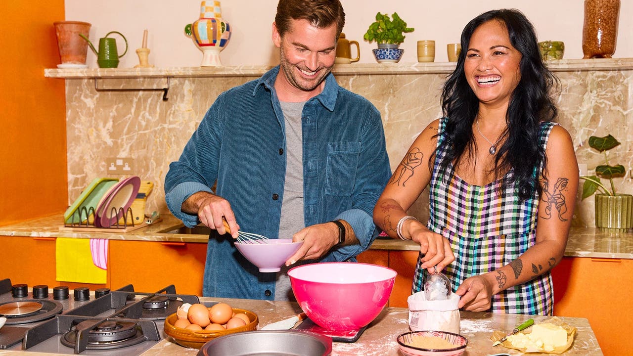 A man and a woman laughing while baking together in a kitchen, surrounded by baking ingredients like eggs and flour.
