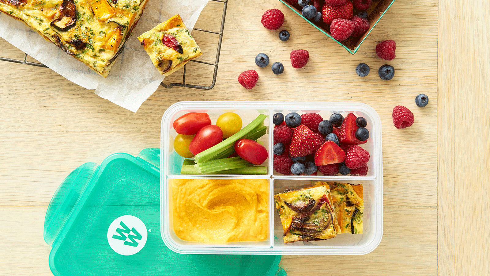 A bento box with tomatoes, celery, mixed berries, hummus, and vegetable frittata. The box has a teal WeightWatchers lid on a wooden table.