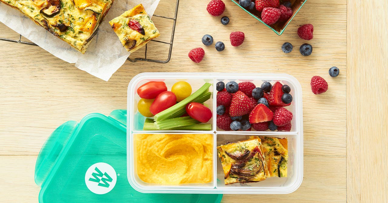 A bento box with tomatoes, celery, mixed berries, hummus, and vegetable frittata. The box has a teal WeightWatchers lid on a wooden table.