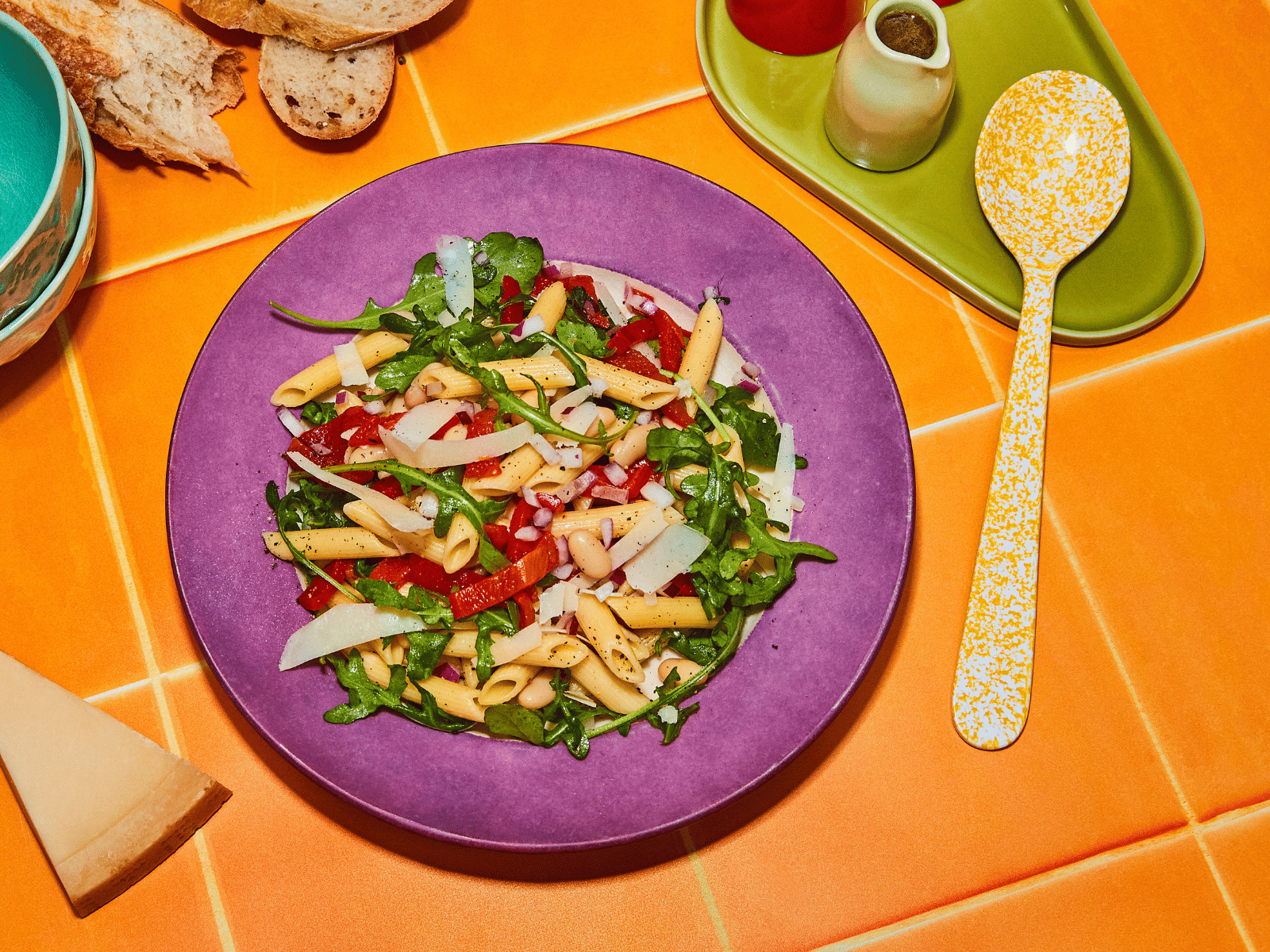 Colourful pasta salad with rocket, capsicum, and shaved Parmesan in a purple bowl on an orange tiled surface, accompanied by sliced bread and condiments.