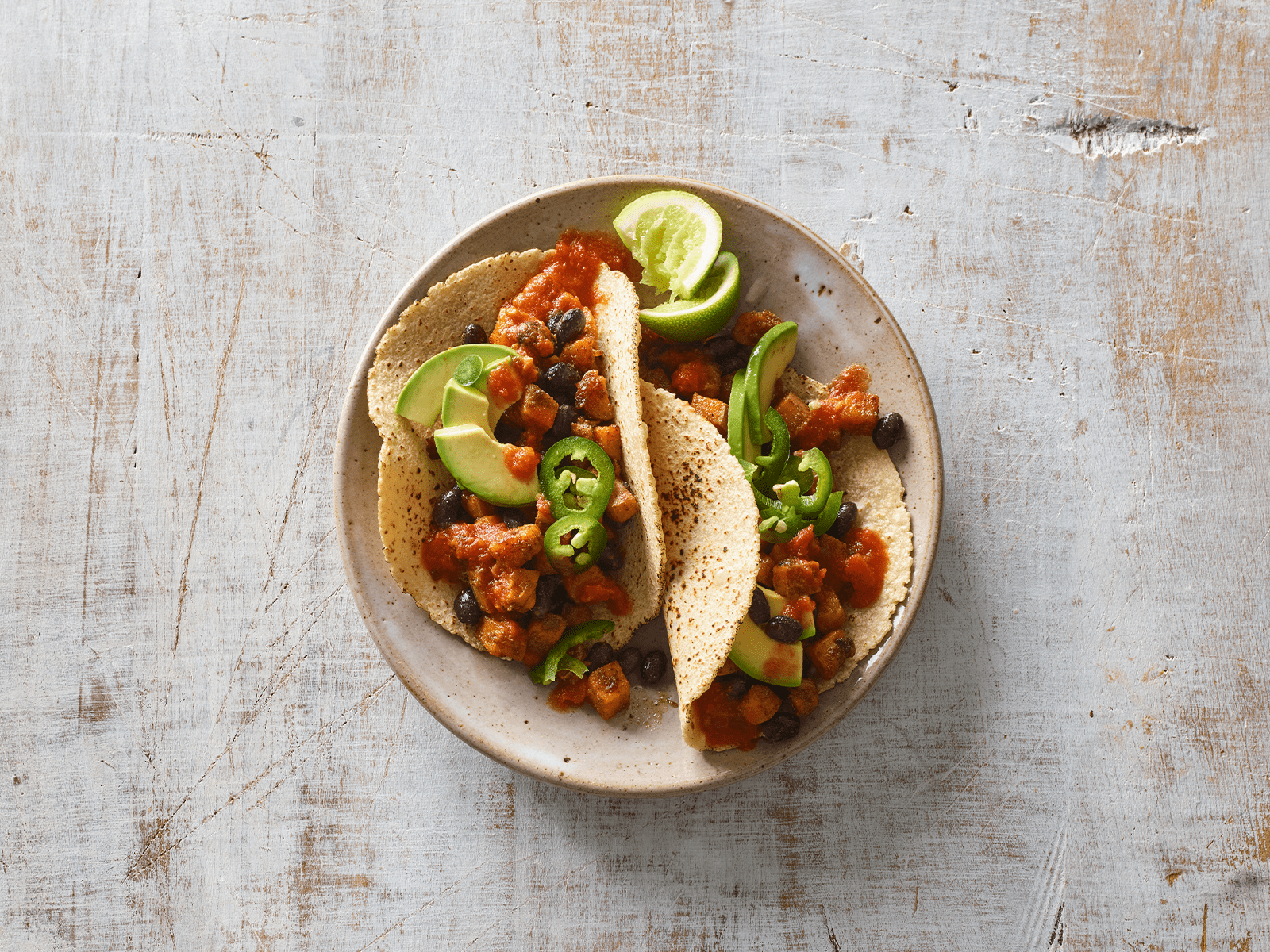 Plate of chicken tacos with black beans, avocado slices, jalapeños, salsa, and lime wedges, served on corn tortillas on a rustic wooden table.