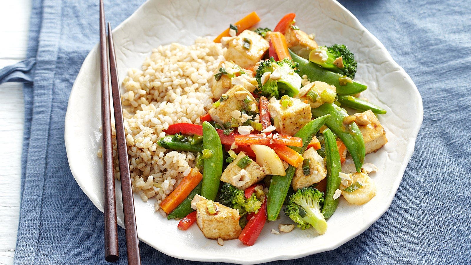 A plate of stir-fried vegetables and tofu served with brown rice, accompanied by chopsticks on a blue cloth.