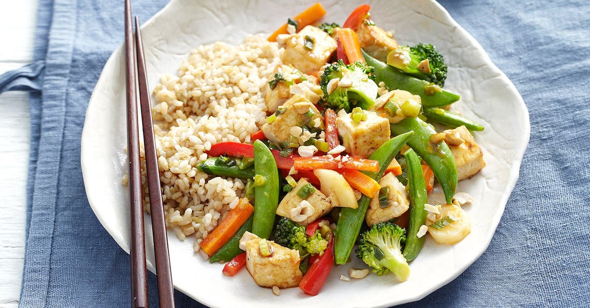 A plate of stir-fried vegetables and tofu served with brown rice, accompanied by chopsticks on a blue cloth.