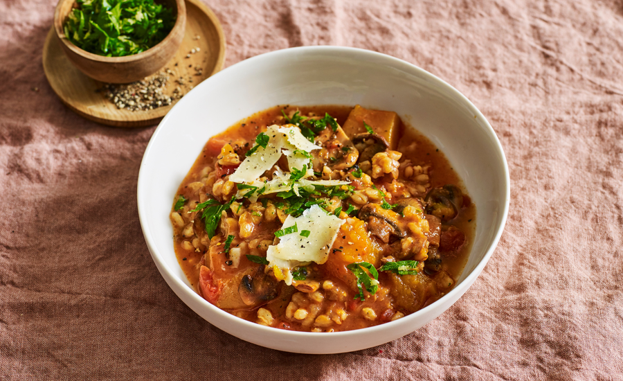 Hearty vegetable and barley stew garnished with fresh parsley and Parmesan shavings, served in a white bowl with a side of chopped herbs and cracked black pepper.