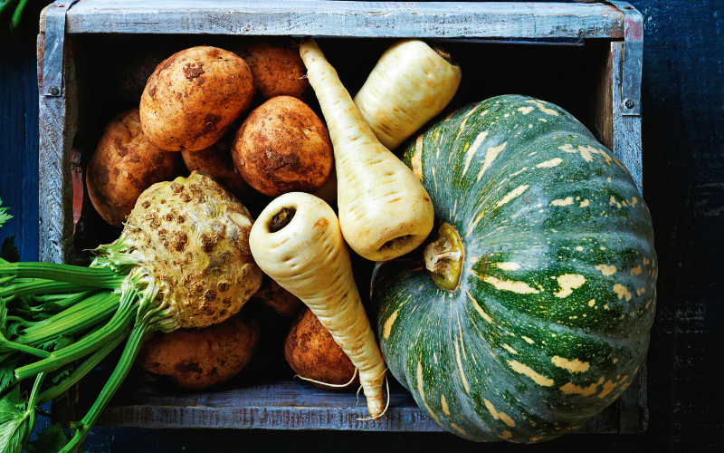 A wooden crate filled with winter root vegetables including potatoes, parsnips, celeriac, and a green pumpkin.