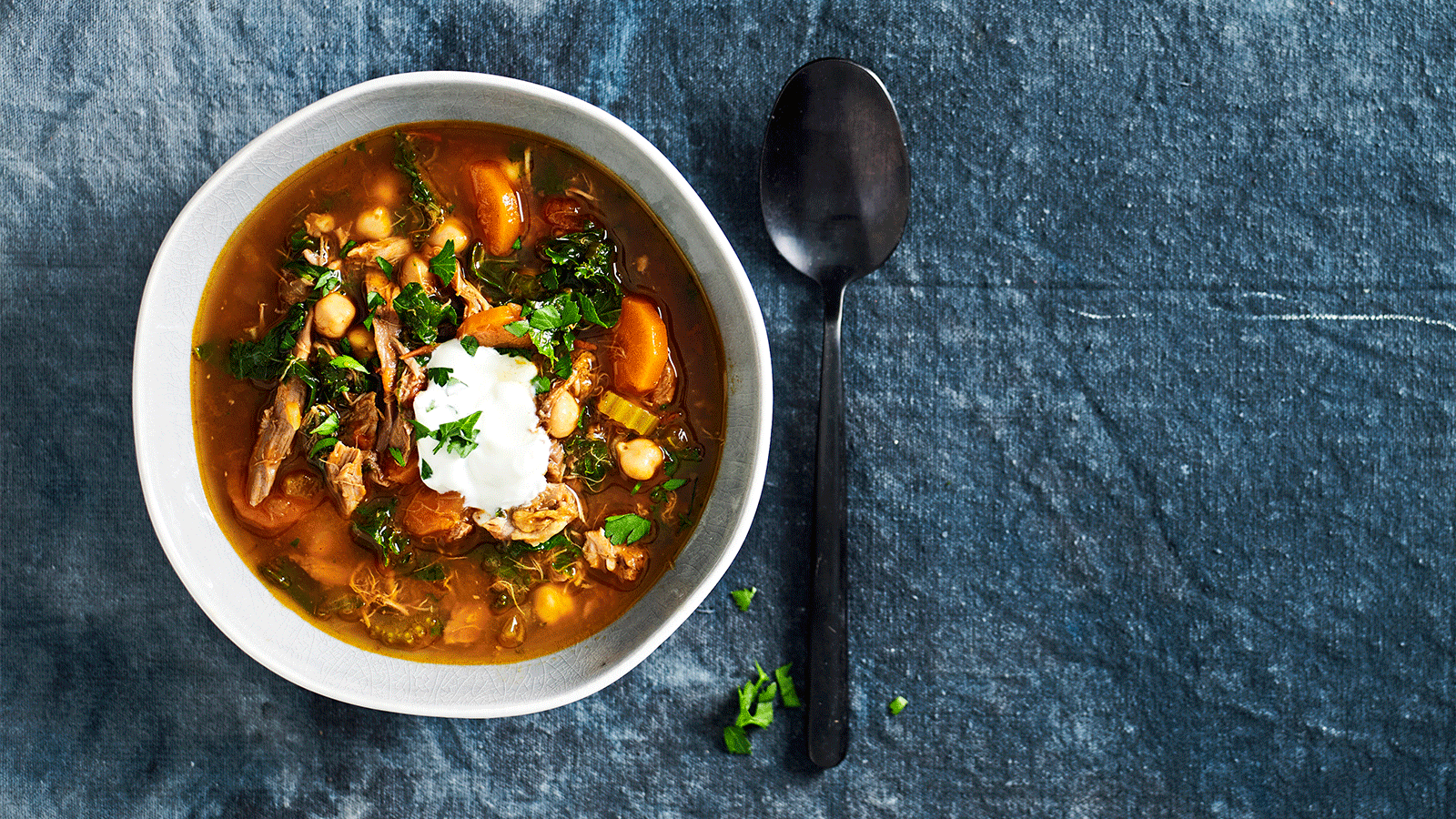 A hearty bowl of vegetable and chickpea soup, garnished with fresh herbs and a dollop of yoghurt, placed on a dark blue textured background.