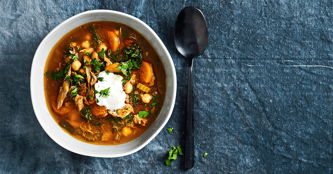 A hearty bowl of vegetable and chickpea soup, garnished with fresh herbs and a dollop of yoghurt, placed on a dark blue textured background.