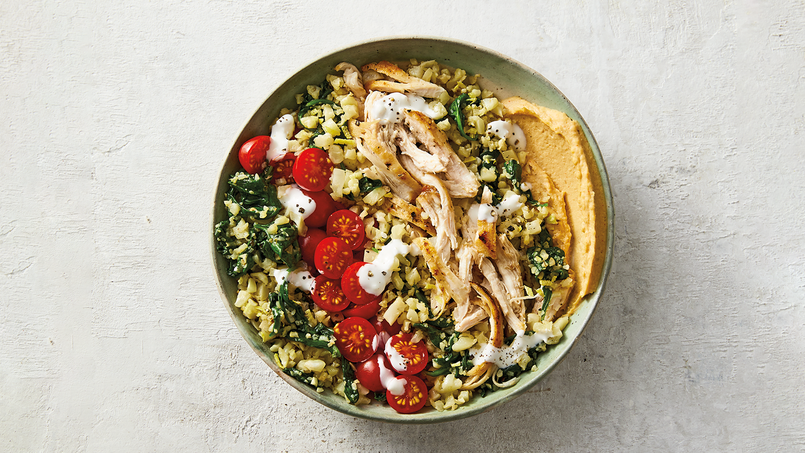A nutritious bowl meal with grilled chicken, cherry tomatoes, greens, grains, and a dollop of hummus.