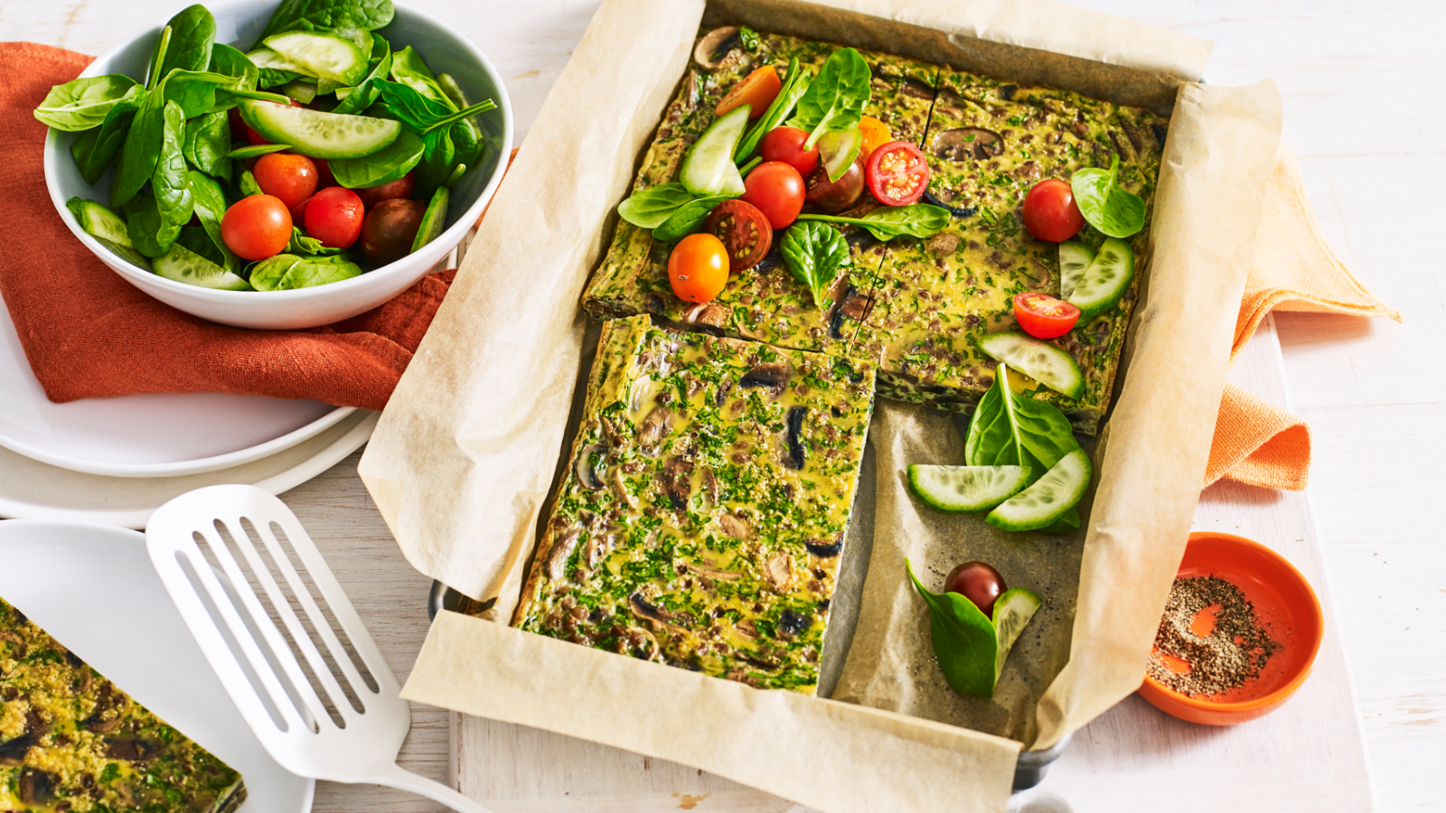 Baked vegetable slice served on parchment paper with fresh spinach, cherry tomatoes, and cucumber salad