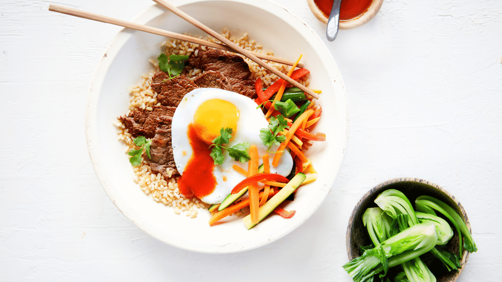 A vibrant bowl of beef served with brown rice, a sunny-side-up egg, and a medley of fresh vegetables garnished with coriander and a drizzle of sauce. Chopsticks rest on the bowl.