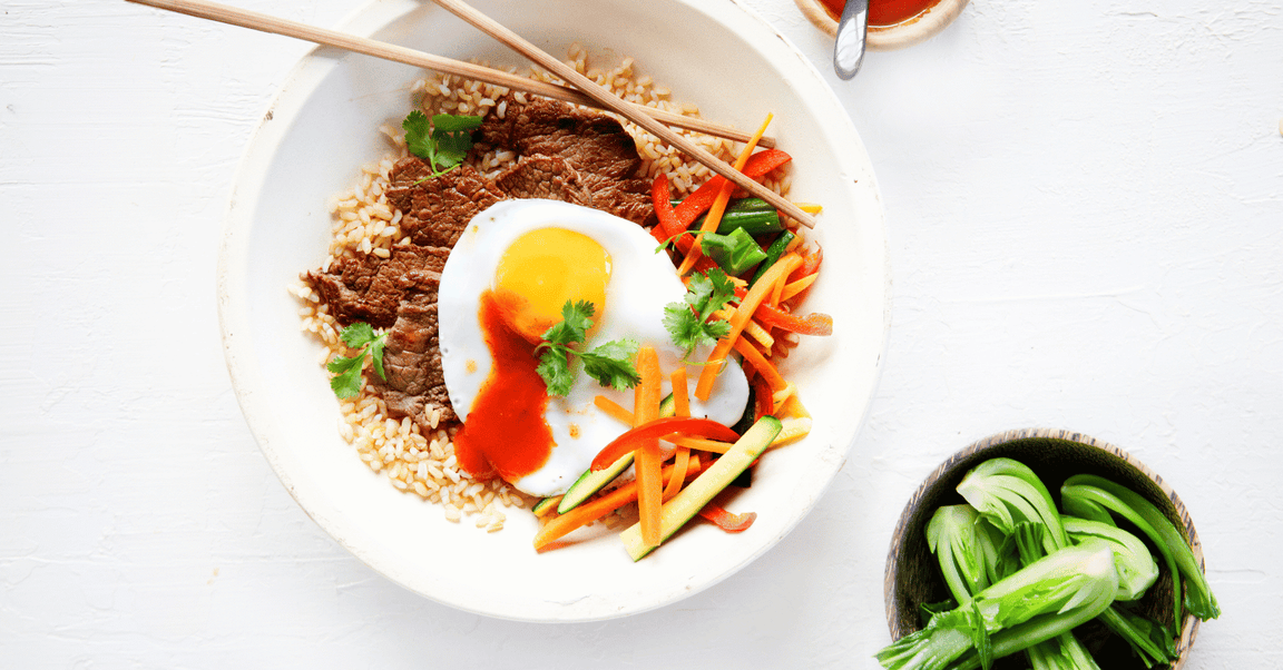 A vibrant bowl of beef served with brown rice, a sunny-side-up egg, and a medley of fresh vegetables garnished with coriander and a drizzle of sauce. Chopsticks rest on the bowl.