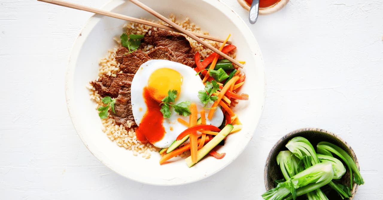 A vibrant bowl of beef served with brown rice, a sunny-side-up egg, and a medley of fresh vegetables garnished with coriander and a drizzle of sauce. Chopsticks rest on the bowl.