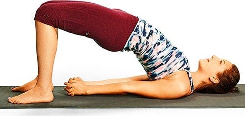 A woman practicing the bridge pose on a yoga mat, wearing maroon leggings and a patterned top.