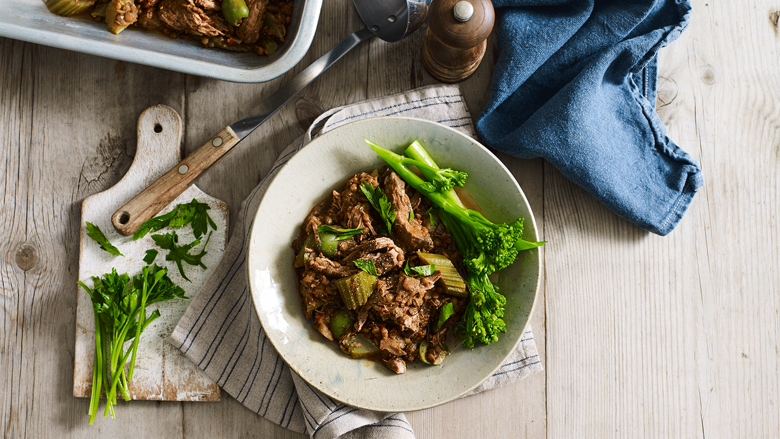A delicious dish with shredded meat, vegetables, and green broccolini on a white plate, garnished with fresh herbs.