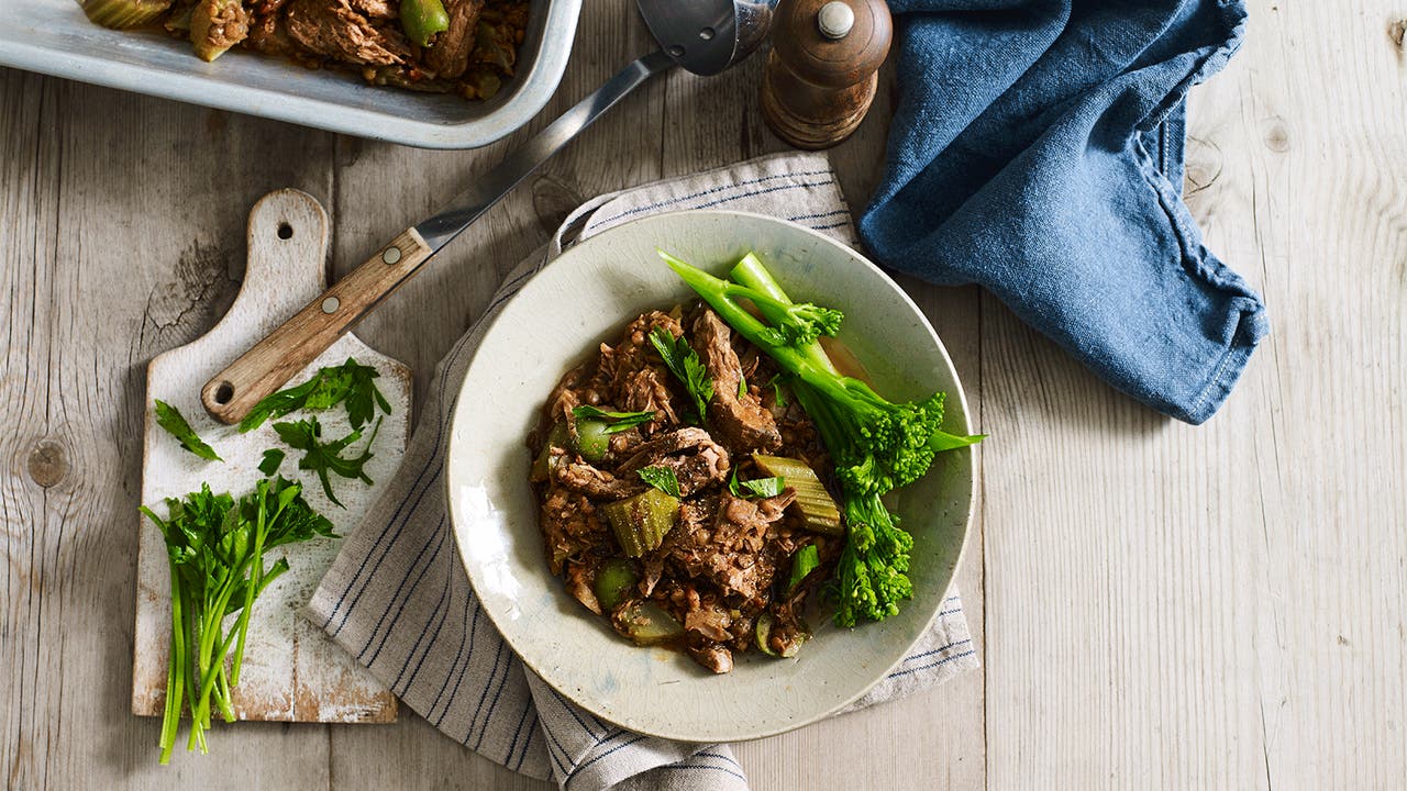 A delicious dish with shredded meat, vegetables, and green broccolini on a white plate, garnished with fresh herbs.