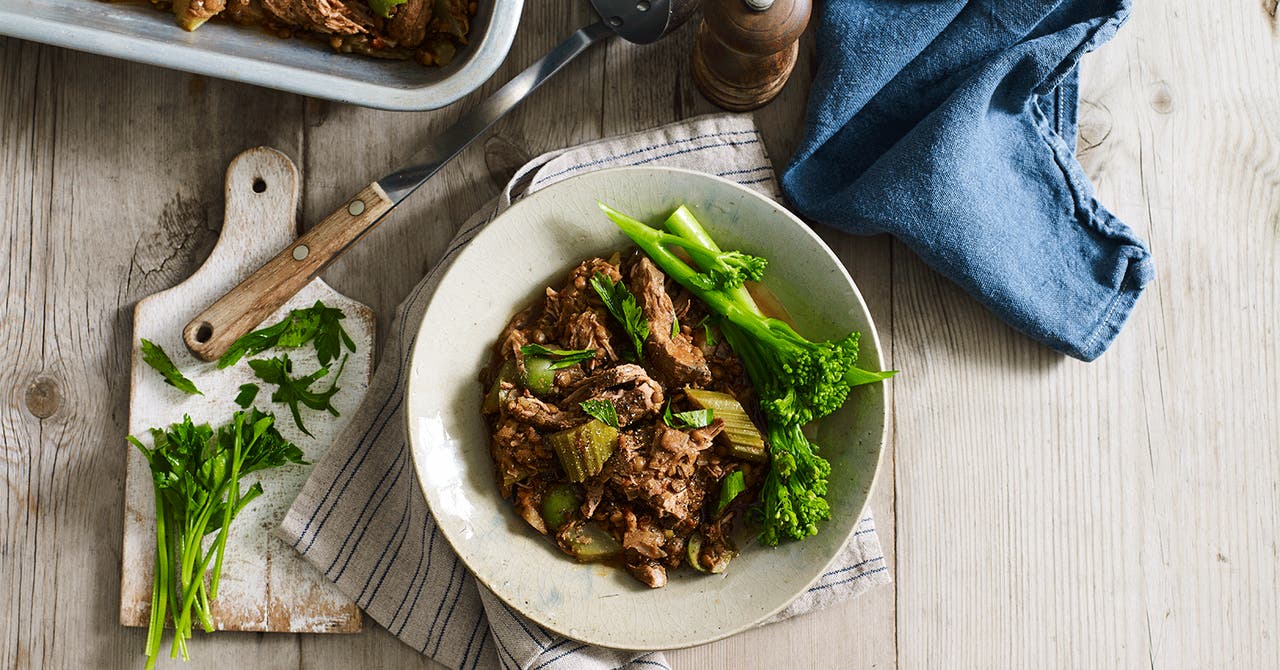A delicious dish with shredded meat, vegetables, and green broccolini on a white plate, garnished with fresh herbs.