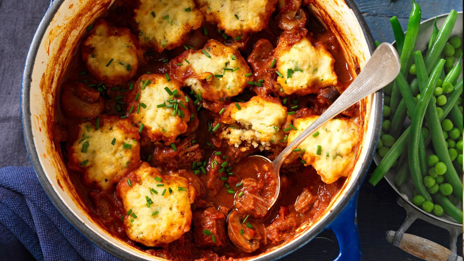 A close-up of a hearty beef stew topped with golden dumplings and garnished with fresh chives, served in a blue cast iron pot with a side of green beans and peas.
