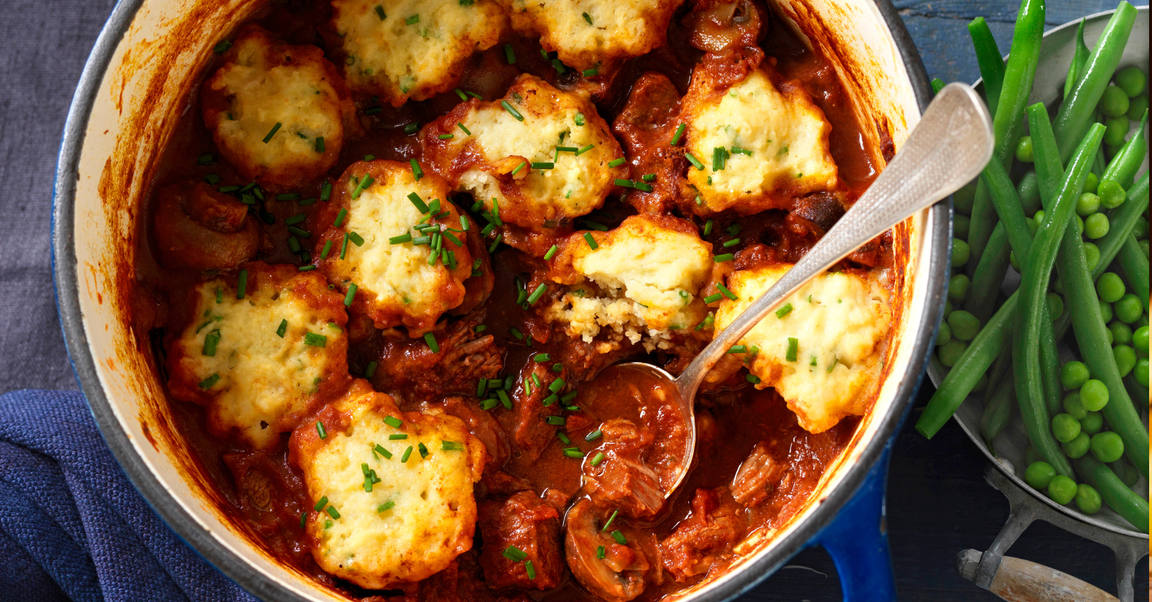 A close-up of a hearty beef stew topped with golden dumplings and garnished with fresh chives, served in a blue cast iron pot with a side of green beans and peas.