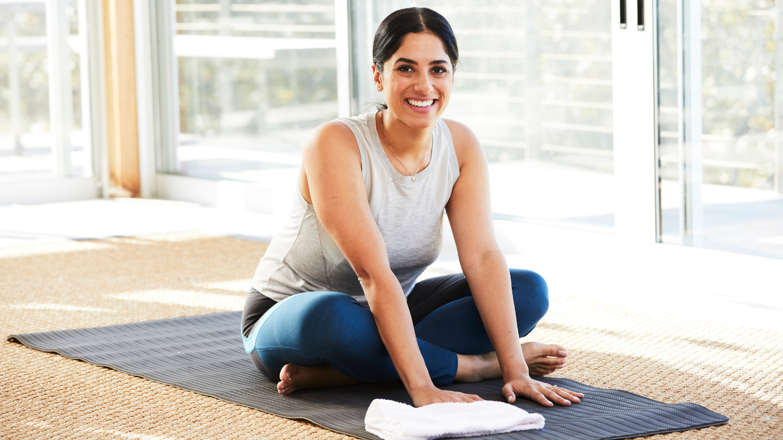 A smiling woman sitting on a yoga mat in a cross-legged position, in a bright room with large windows.