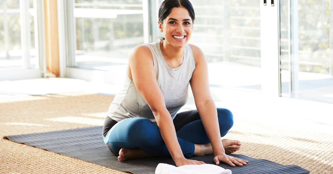 A smiling woman sitting on a yoga mat in a cross-legged position, in a bright room with large windows.