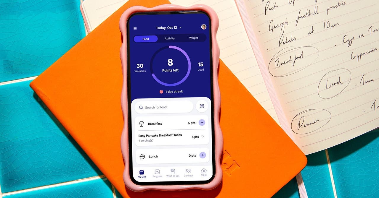 Close-up of a smartphone with the WeightWatchers app open, showing daily points tracking, placed on an orange notebook with handwritten notes in the background.