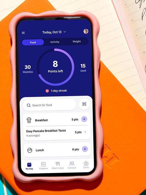 Close-up of a smartphone with the WeightWatchers app open, showing daily points tracking, placed on an orange notebook with handwritten notes in the background.