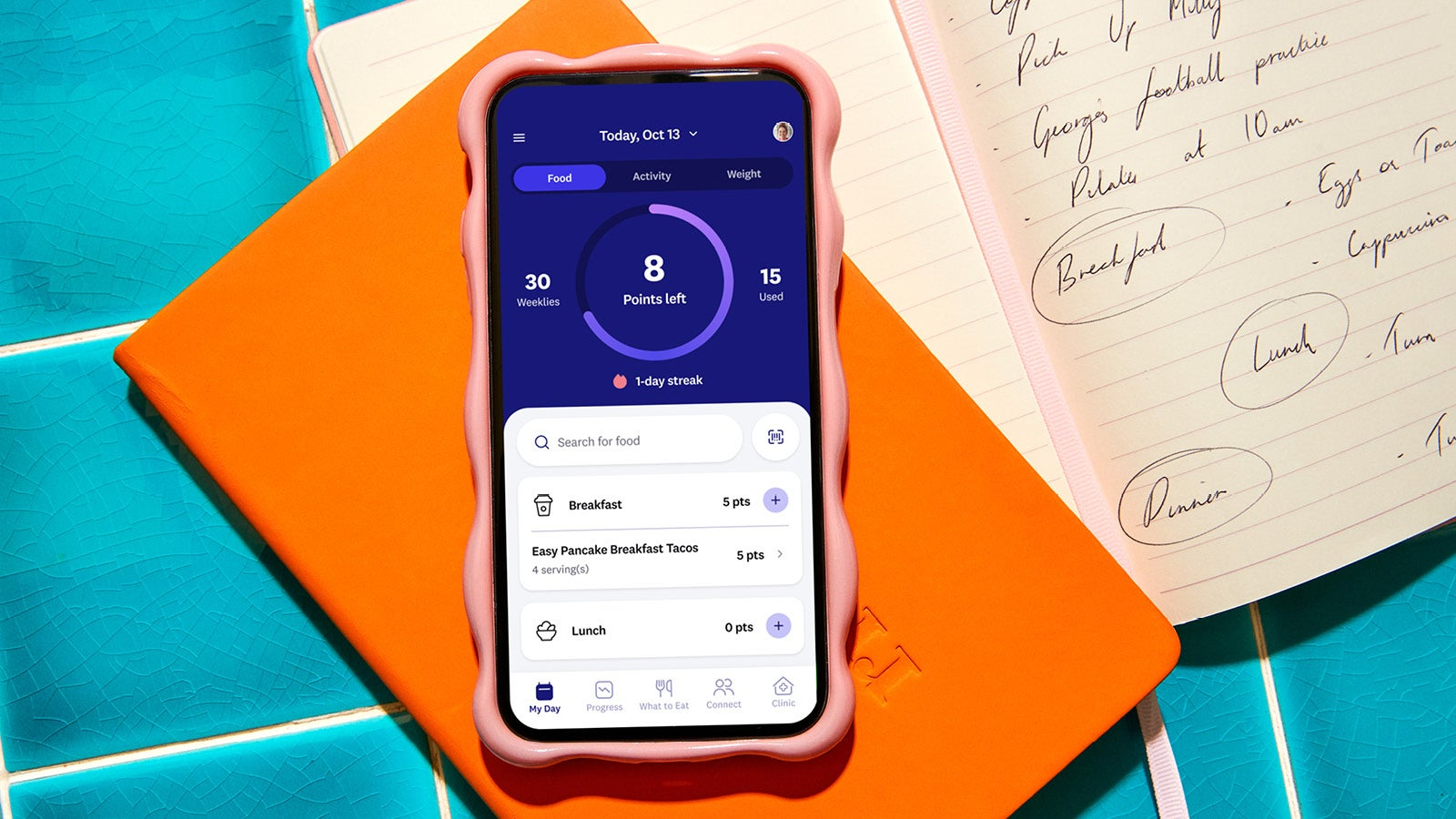 Un téléphone intelligent affichant une application de suivi alimentaire sur un cahier orange avec des notes manuscrites à proximité.