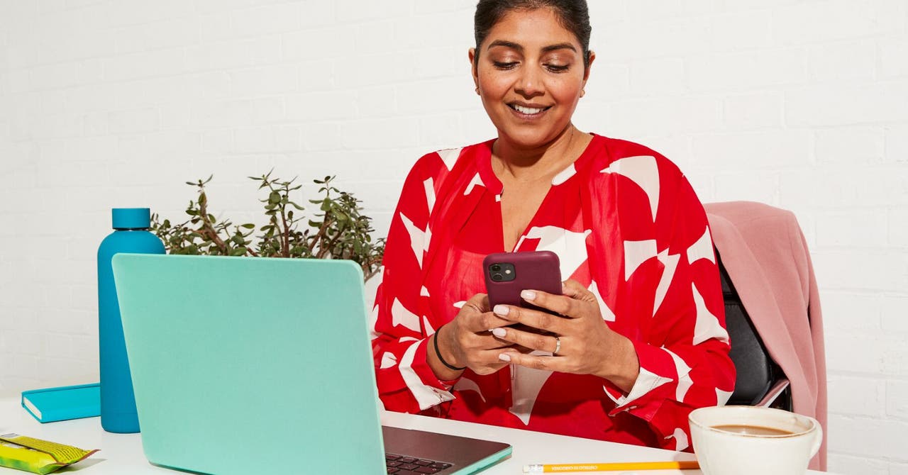 Une femme assise à un bureau utilisant un téléphone intelligent, avec un portable, un café et des collations à proximité.
