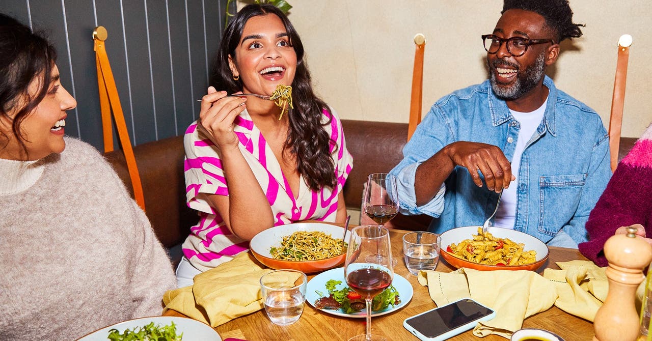 Un groupe d'amis profitant d'un repas ensemble au restaurant, avec une femme riant et tenant une fourchette de pâtes.