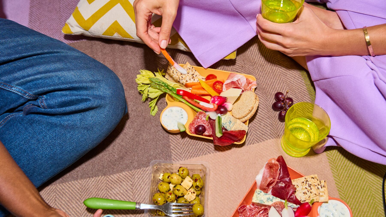Une vue rapprochée d'un pique-nique avec deux personnes savourant un plateau coloré de collations, incluant des légumes, du fromage, et des viandes séchées.