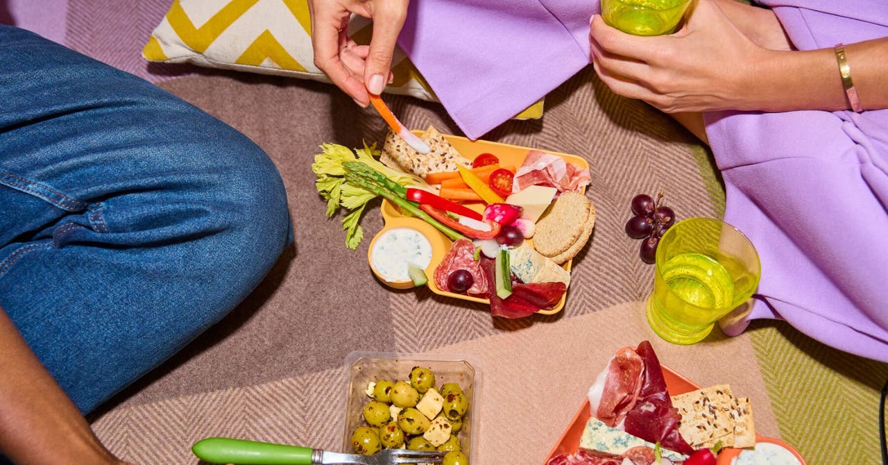 Une vue rapprochée d'un pique-nique avec deux personnes savourant un plateau coloré de collations, incluant des légumes, du fromage, et des viandes séchées.