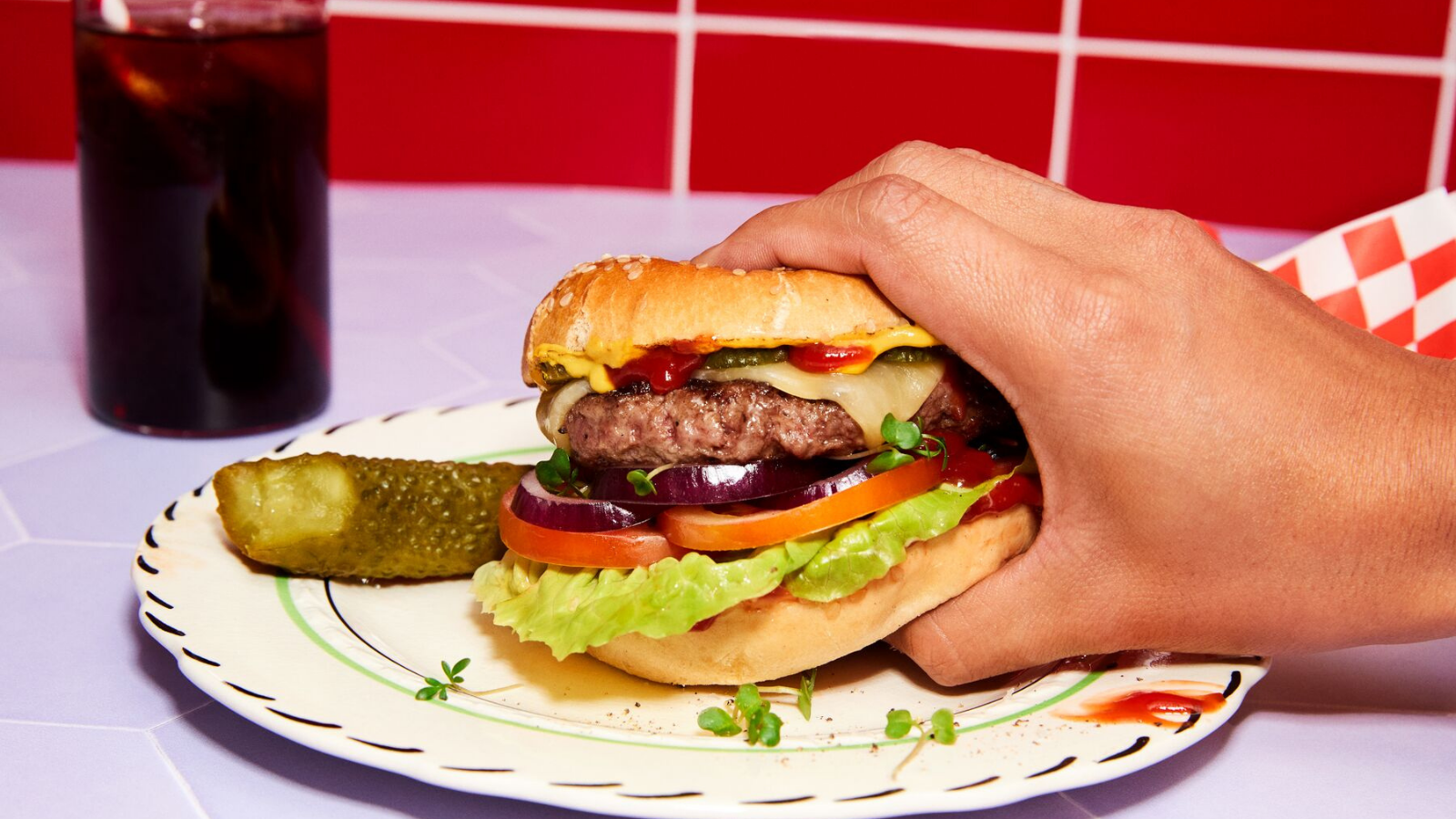 Gros plan d'une main tenant un burger avec laitue, tomate, fromage et cornichons, avec un cornichon à côté.