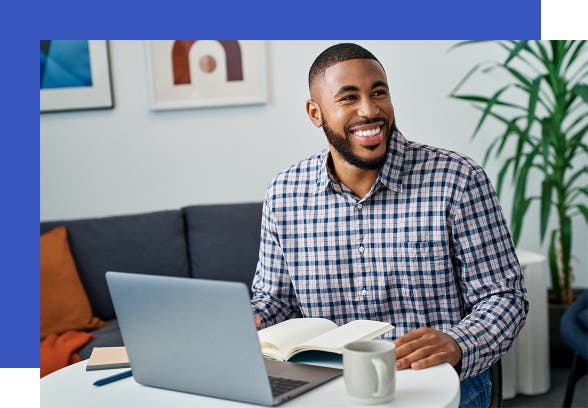A black man sits at a table with his laptop and a coffee.